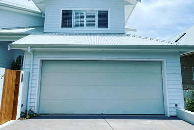 White sectional garage door on a two-storey coastal-style weatherboard home, with a timber side gate and steel roof above.