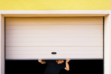 Person's hands lifting an off-white sectional garage door from underneath on a yellow building facade