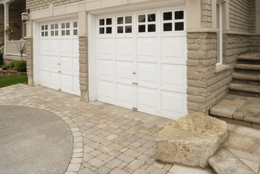 Two white raised-panel garage doors with windowed upper panels on a stone-and-brick house with a paver driveway