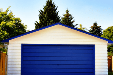 Blue sectional garage door on a white garage building with a pitched roof and trees behind