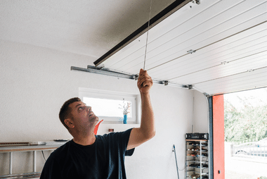 Man reaching up to pull the release cord on a white sectional garage door track inside a garage