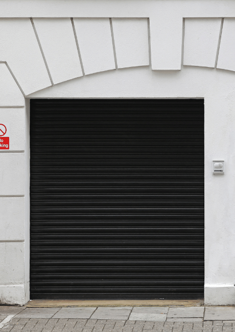 Black roller door in a white archway with a no-parking sign and intercom panel beside it