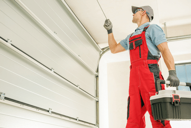 Tradesman in red overalls holding a toolbox reaching up to a white sectional garage door track