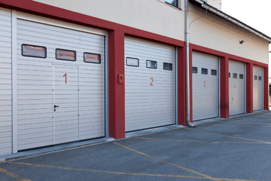 Row of white sectional garage doors with red trim and numbering on a commercial building.