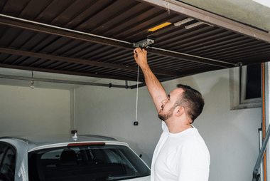Man reaching up to adjust an overhead garage door opener rail inside a home garage, with a car parked below.