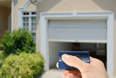 Hand pointing a garage door remote at a beige house's open roller door