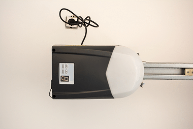 Close-up of a garage door opener motor unit mounted on the ceiling rail with its power cord hanging beside it.