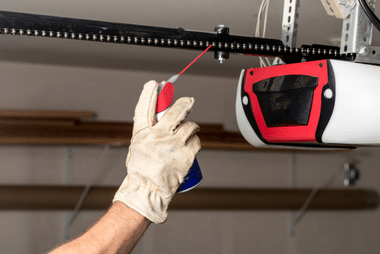Gloved hand applying lubricant spray to a chain-drive garage door opener rail.
