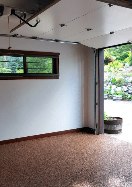 Interior of a renovated home garage with a pebble-finish epoxy floor, a small window, and the door open to a garden.