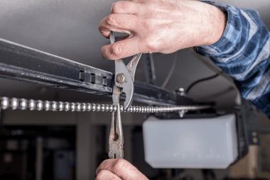Close-up of hands using pliers on a cable and spring assembly at the top of a garage door track.