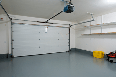 Interior of an empty home garage with a white sectional door, ceiling-mounted opener, and storage shelving to the side.