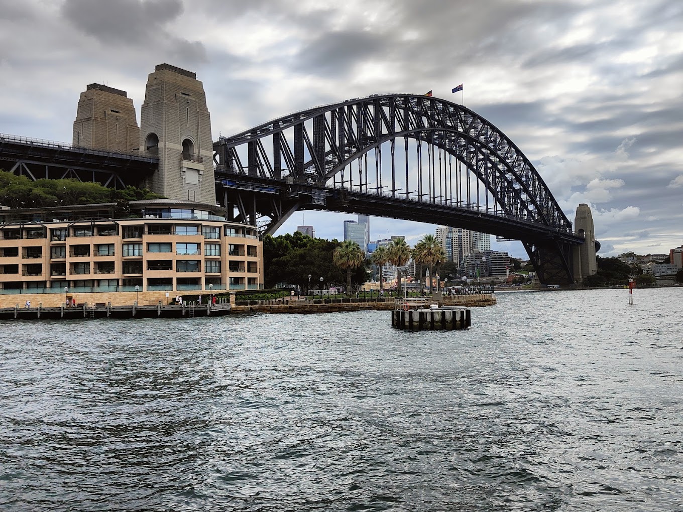 View of the Sydney Harbour Bridge and the city skyline from the water, with a curved waterfront apartment building in the foreground.