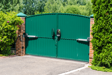 Green vertical-panel driveway gate fitted with a dual-arm automatic gate opener, flanked by brick pillars and hedging.