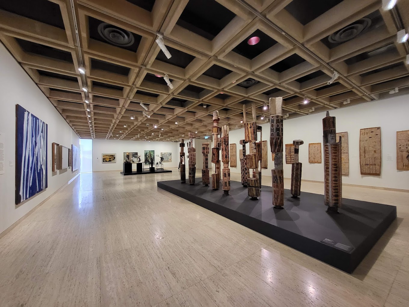 Gallery interior displaying a group of tall carved and painted memorial poles on a low platform, with framed artworks on the surrounding walls.