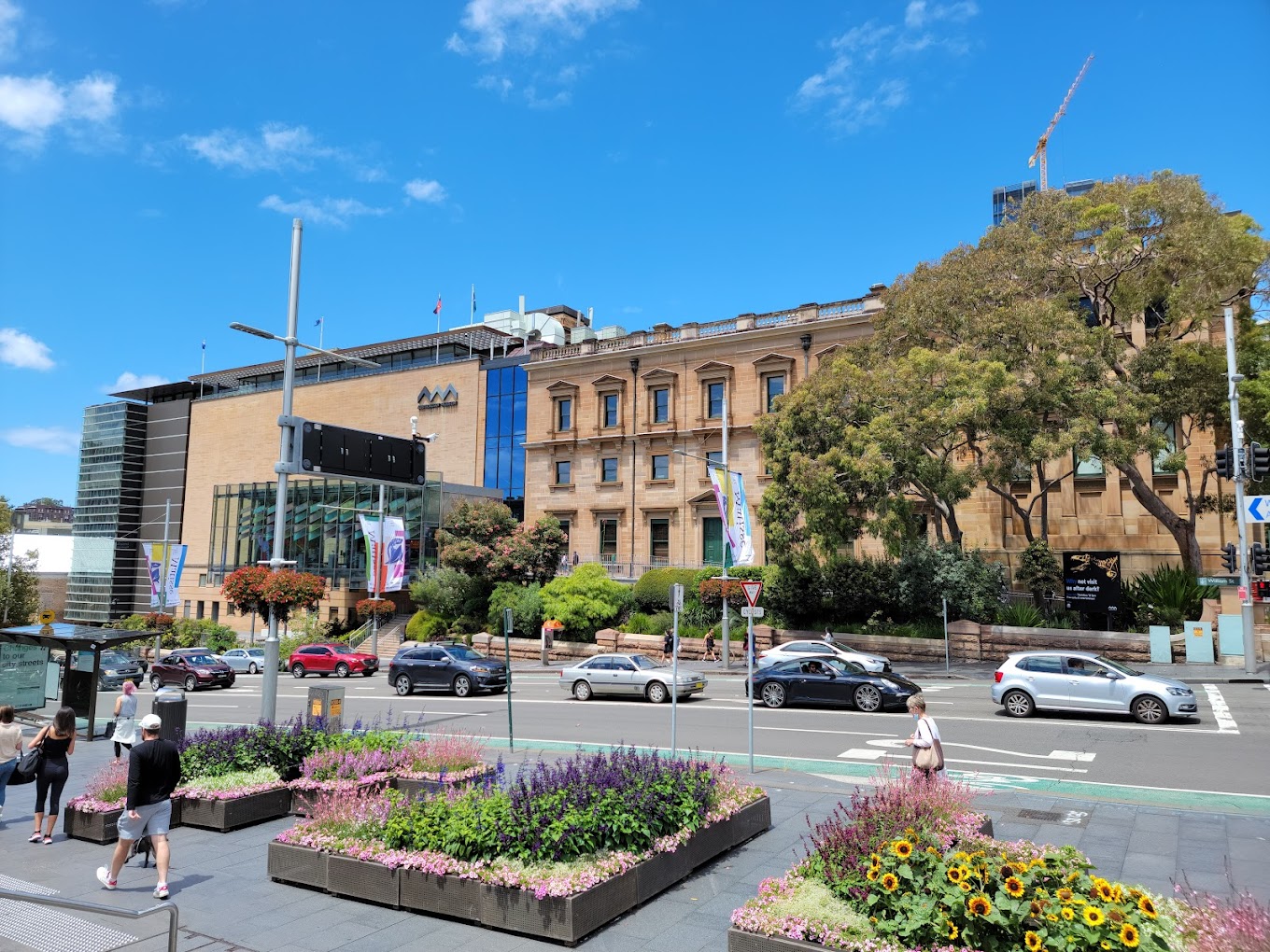 Street view of a sandstone heritage building beside a modern glass extension, with garden beds, banners, and traffic passing in front.