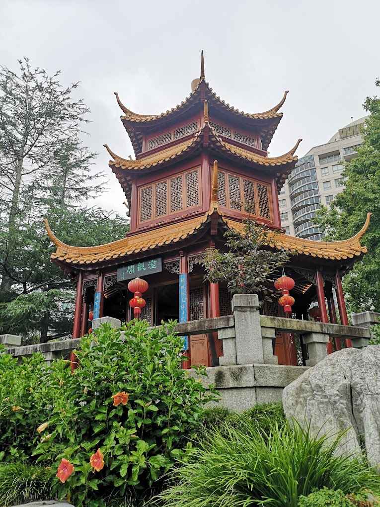 Ornate red and gold multi-tiered pagoda surrounded by garden greenery, with city towers visible behind.