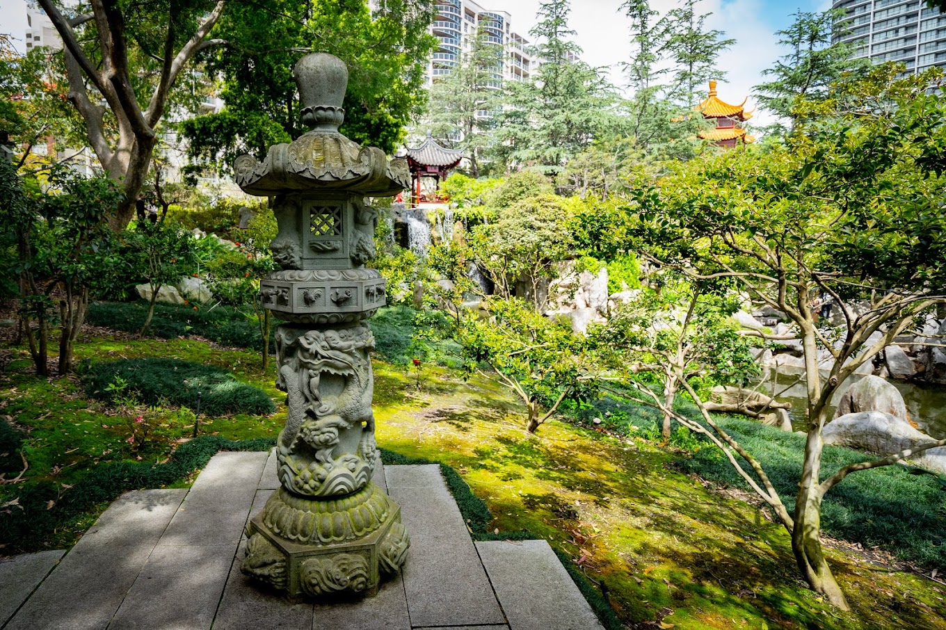 Carved stone lantern in a landscaped garden, with a pagoda and city apartment towers in the background.