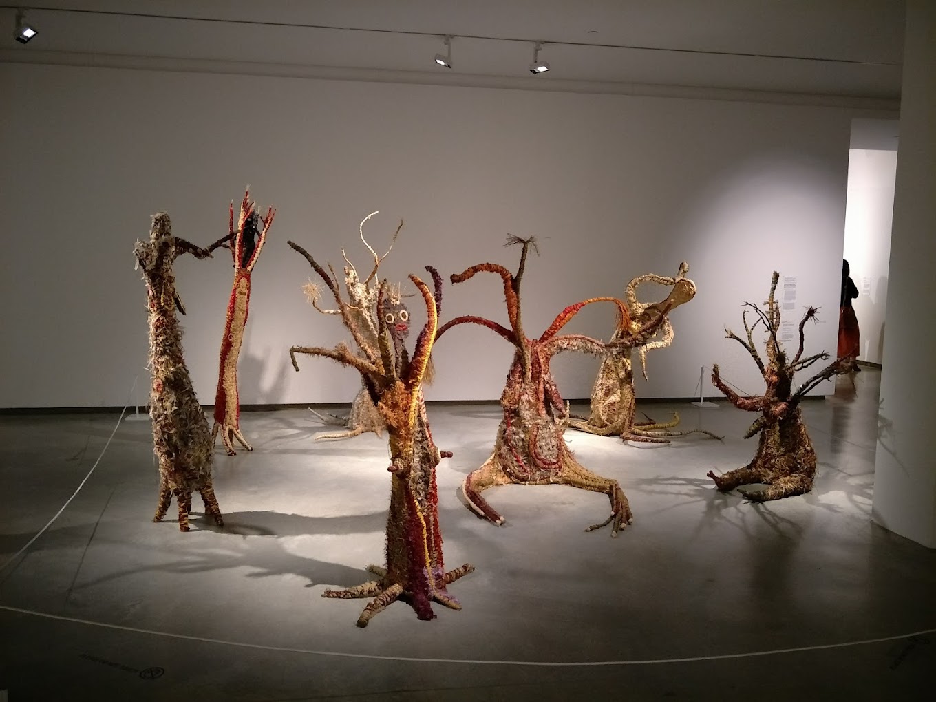 Gallery room displaying several tall sculptures made from woven and dyed natural fibres resembling tree-like figures.