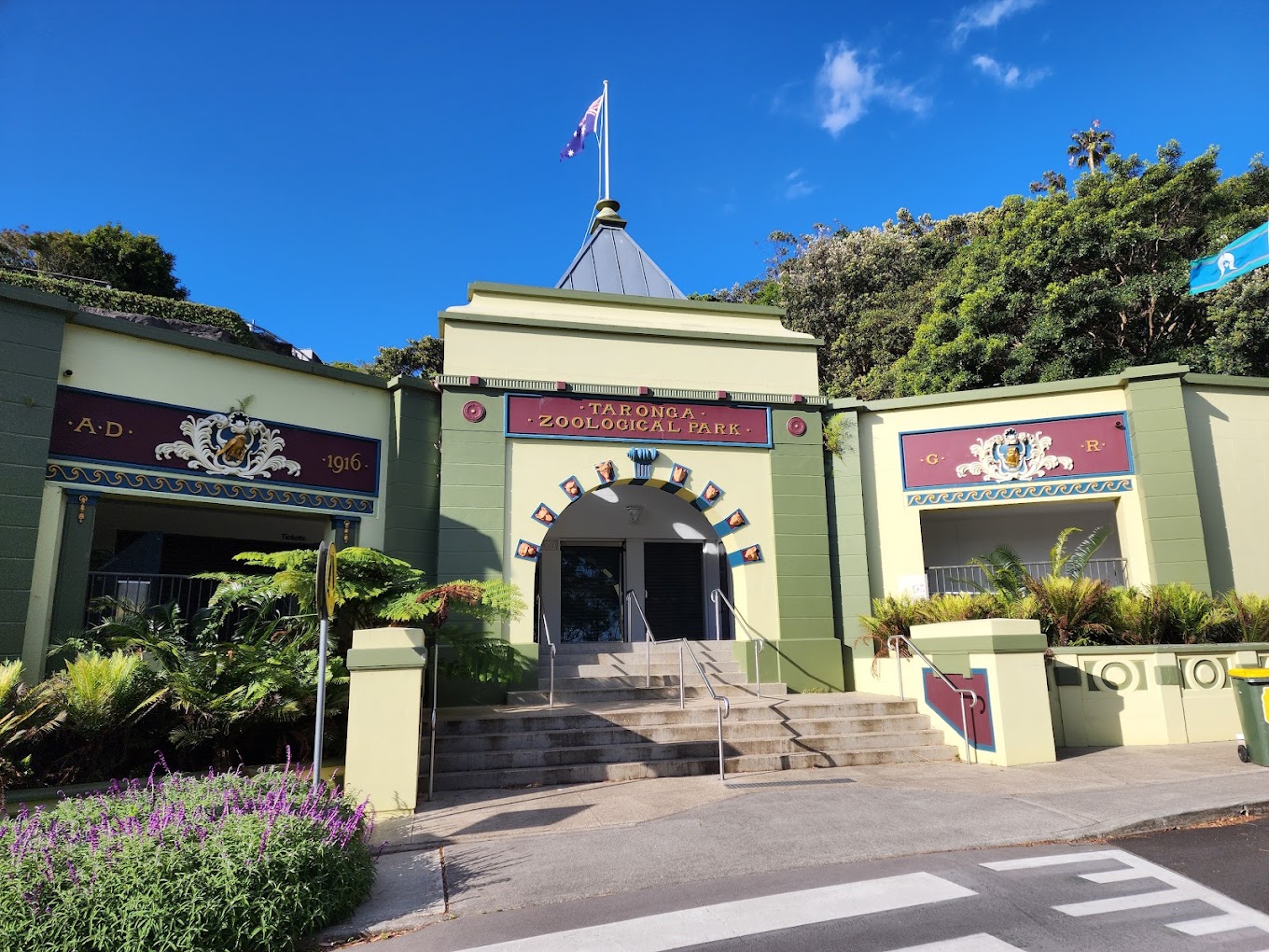 Historic entrance building with 'Taronga Zoological Park' signage above an archway, flanked by garden greenery.