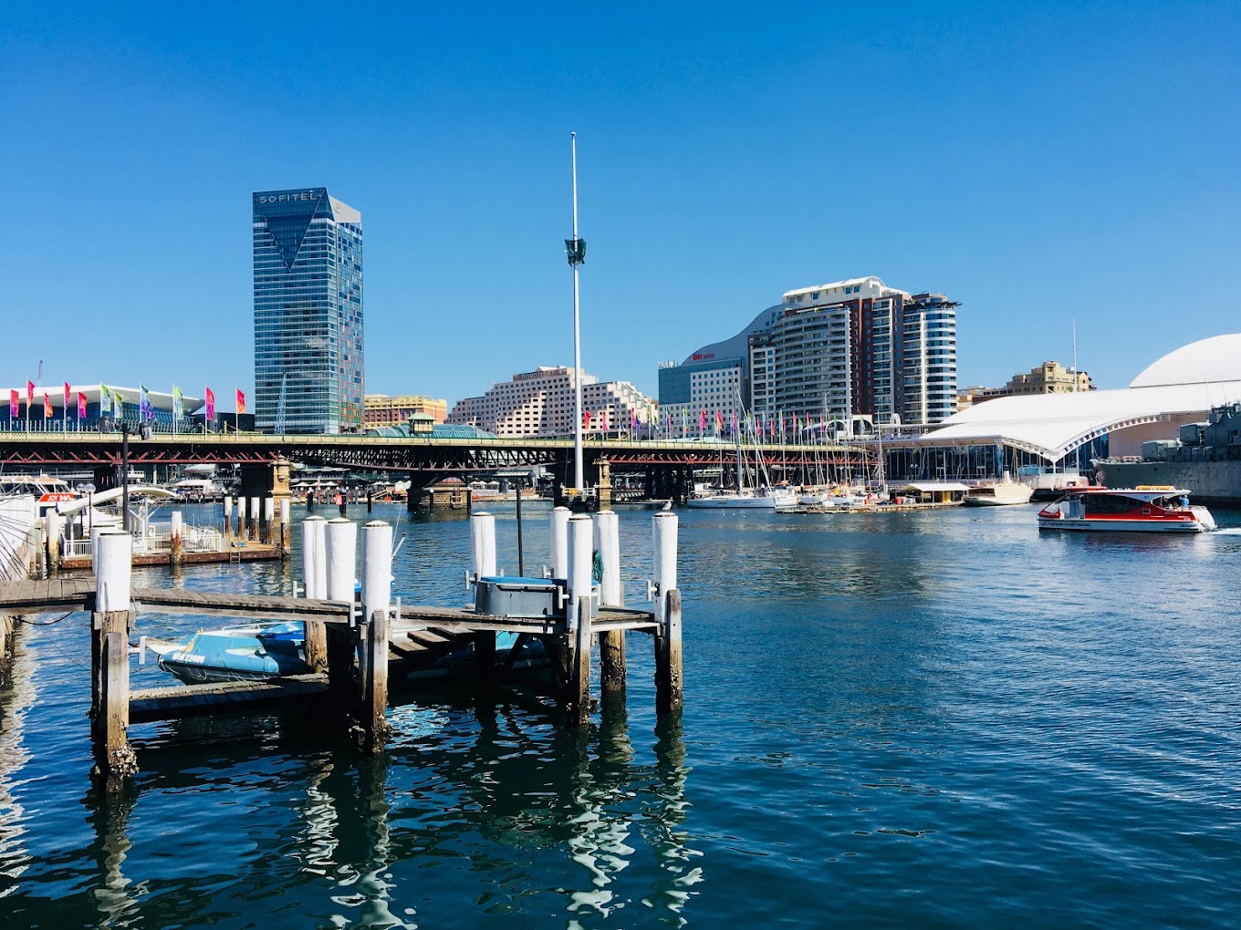 Darling Harbour waterfront with a tall glass hotel tower, apartment buildings, and boats moored along the pier.
