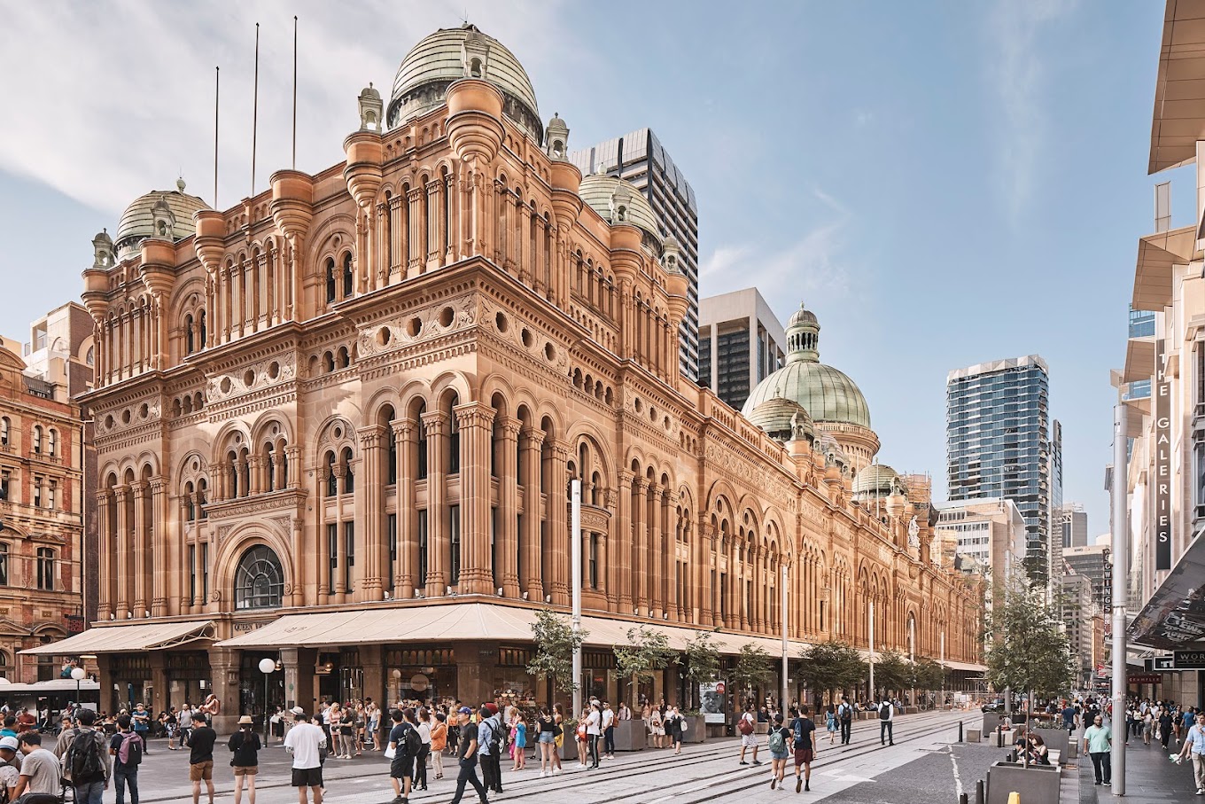 Ornate sandstone facade of the Queen Victoria Building with its domed towers, seen from a busy street with pedestrians.