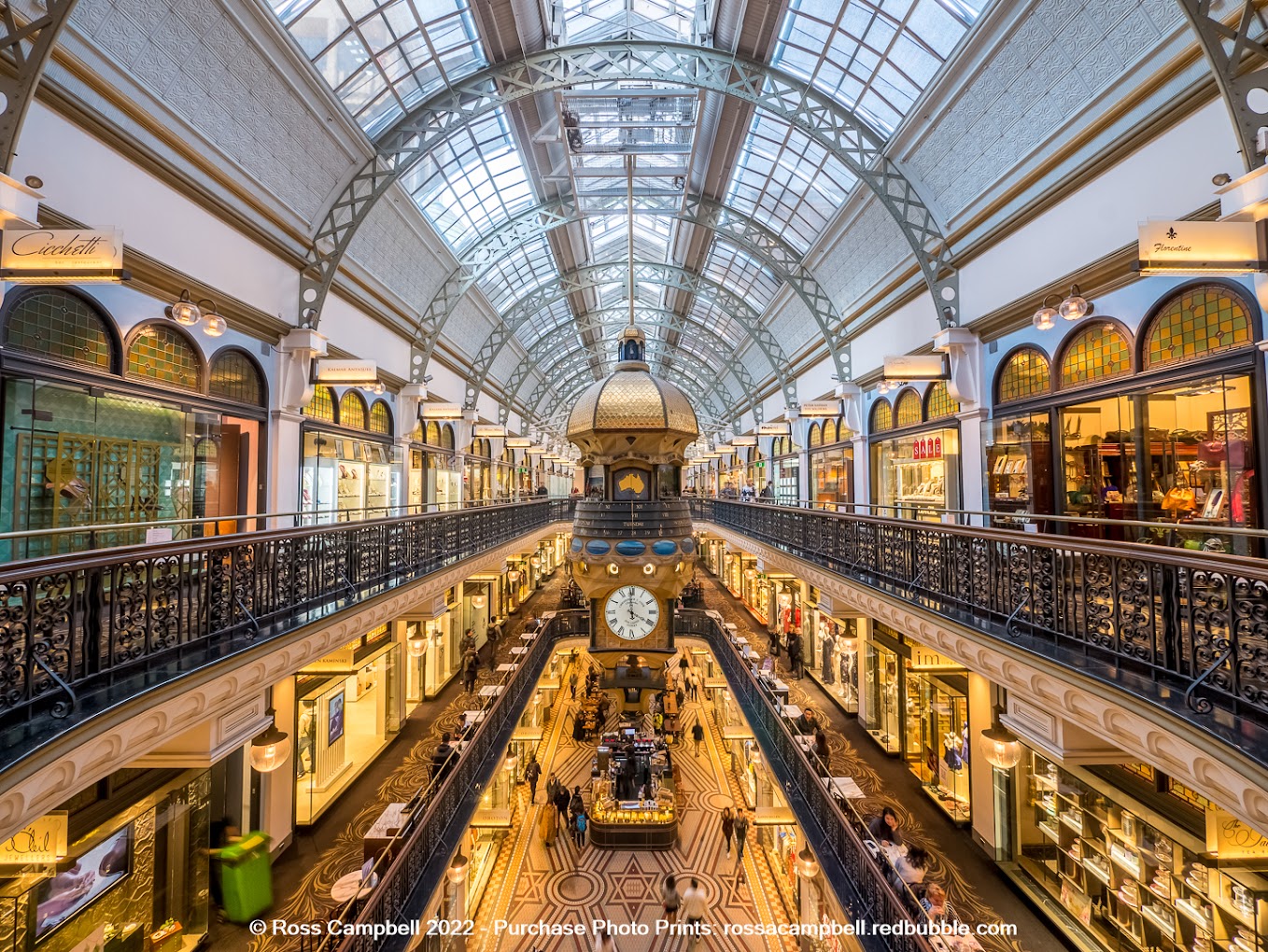 Interior of the Queen Victoria Building arcade, showing its glass barrel-vaulted ceiling and the large hanging clock above shopfronts.