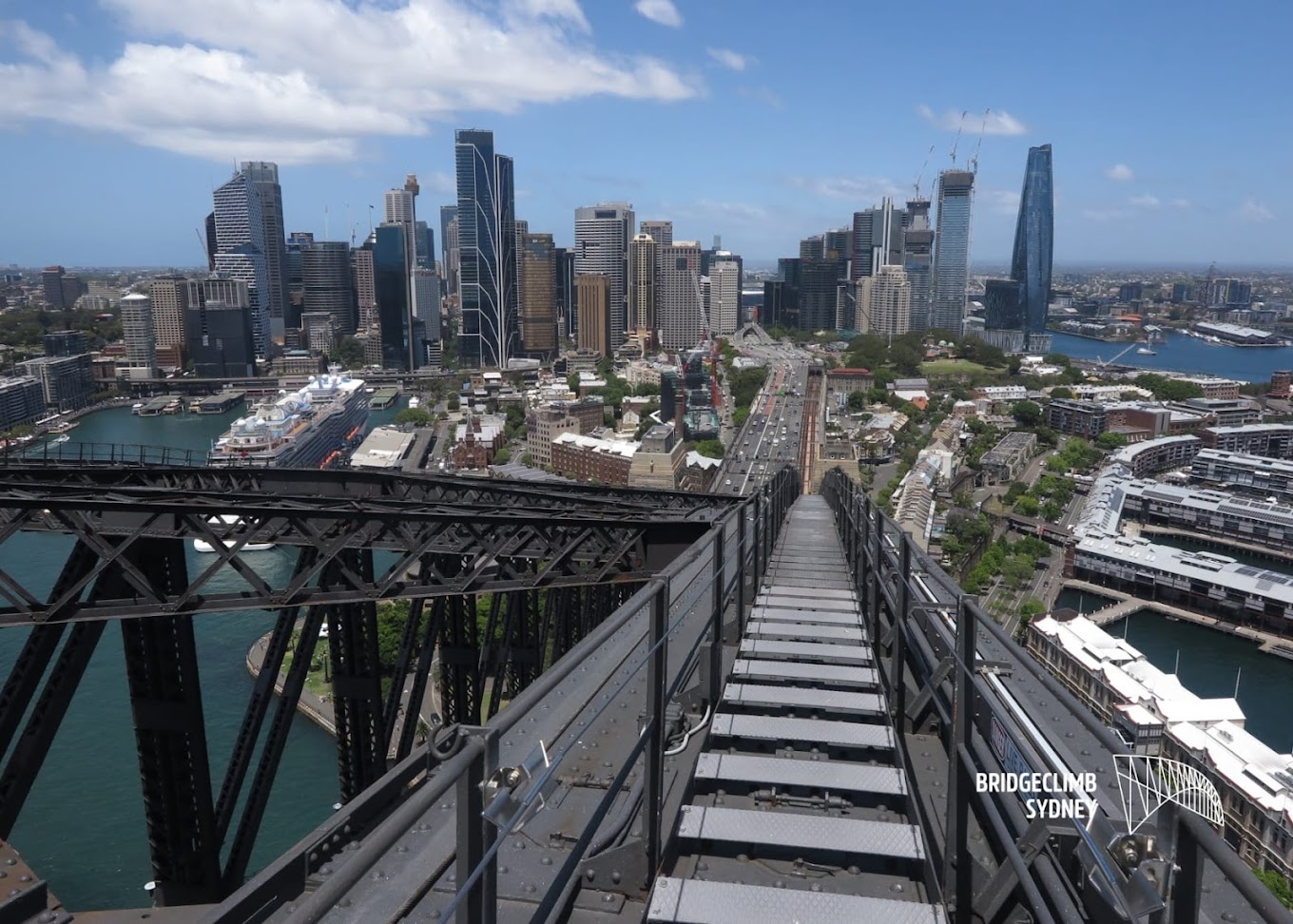 Aerial view from the Sydney Harbour Bridge climb walkway looking down toward the city skyline, with a 'Bridgeclimb Sydney' logo overlay.