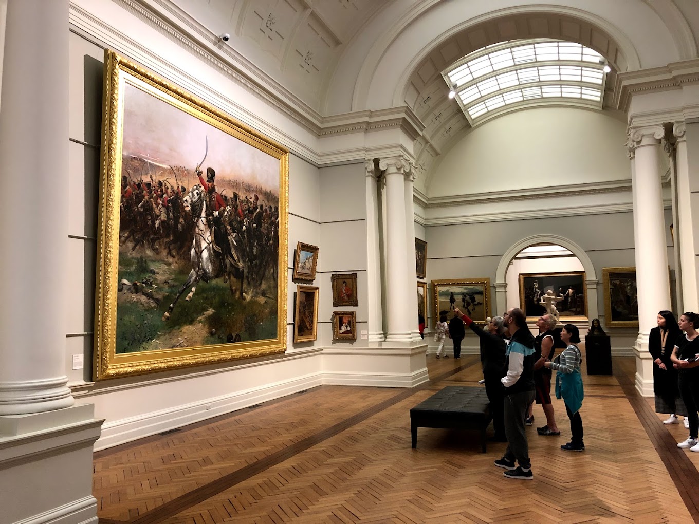 Vaulted gallery room hung with large gilt-framed oil paintings, including a battle scene, with visitors viewing the artwork on a wood floor.